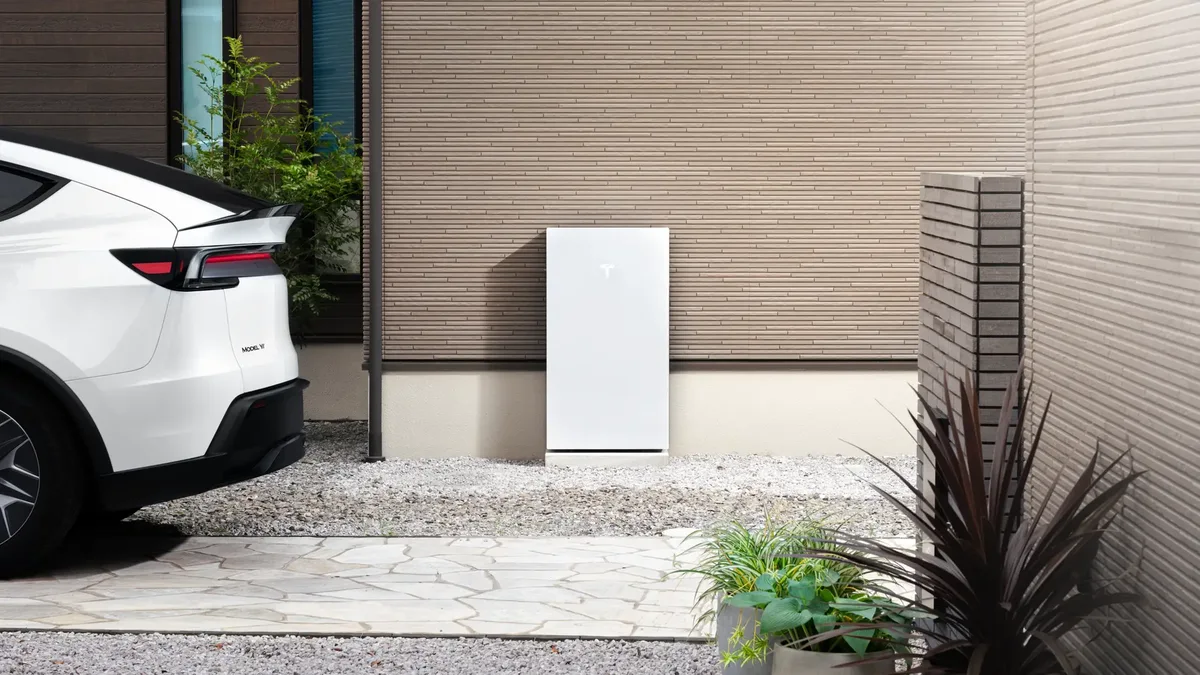 Parte trasera de un Tesla Model Y blanco estacionado junto a una batería doméstica Powerwall instalada en la pared exterior de una vivienda.