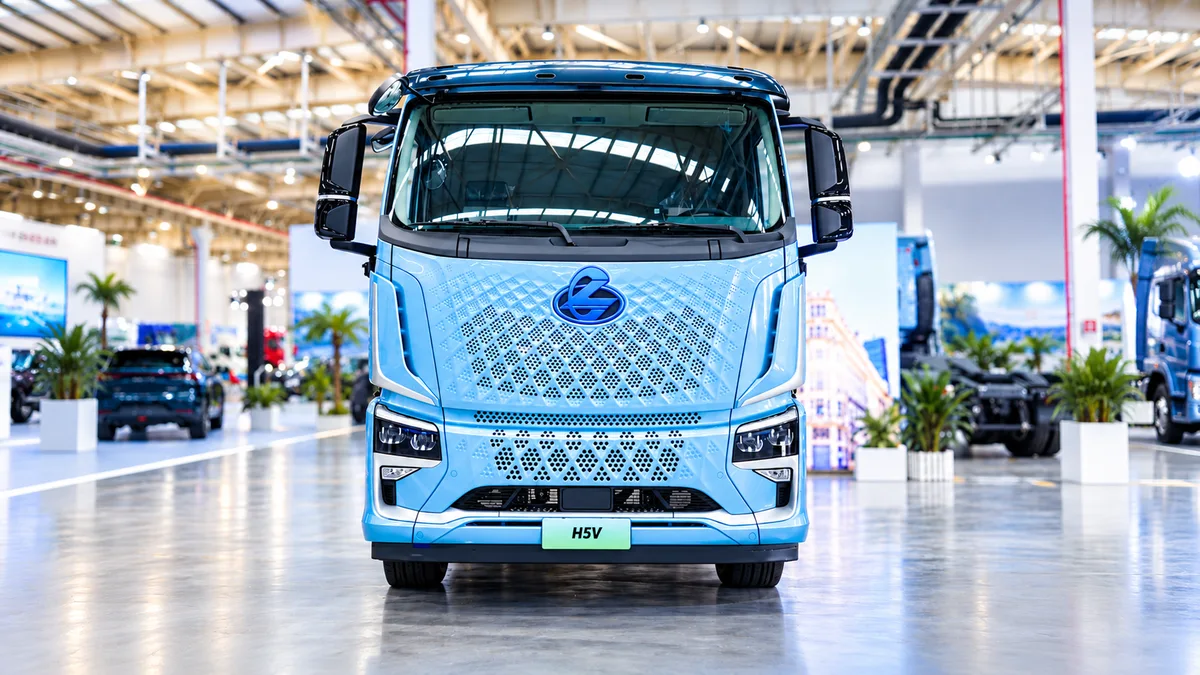 Vista frontal del camión Dongfeng Chenglong H5V azul, con emblema central, parrilla perforada y grandes retrovisores, en un recinto de exposición desenfocado.