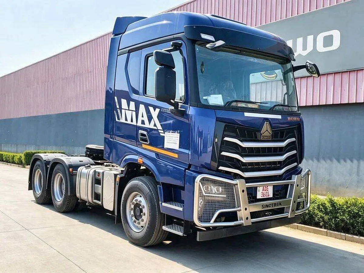 Cabeza tractora azul Sinotruk HOWO MAX de tres ejes, fotografiada de frente y de lado junto a una nave industrial.