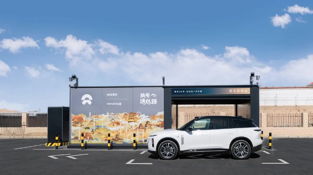 Automóvil eléctrico NIO blanco estacionado ante una estación de intercambio de baterías, con instalaciones modulares y paisaje árido al fondo.