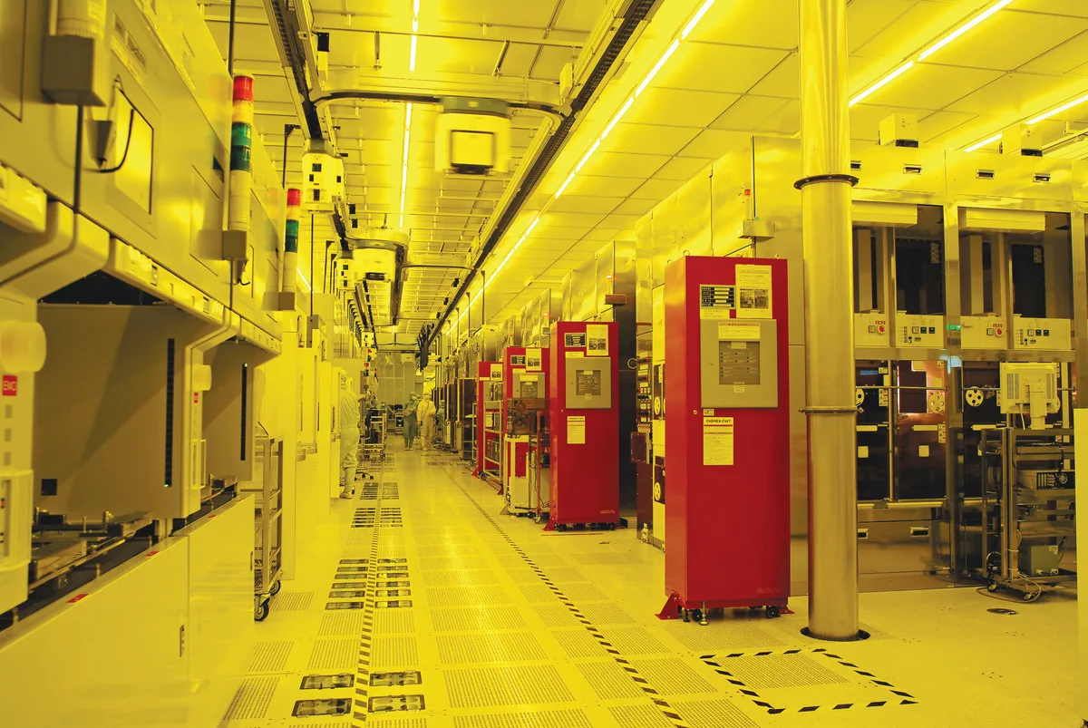 Interior de una sala de fabricación de semiconductores iluminada en amarillo, con maquinaria industrial a ambos lados y personal al fondo.