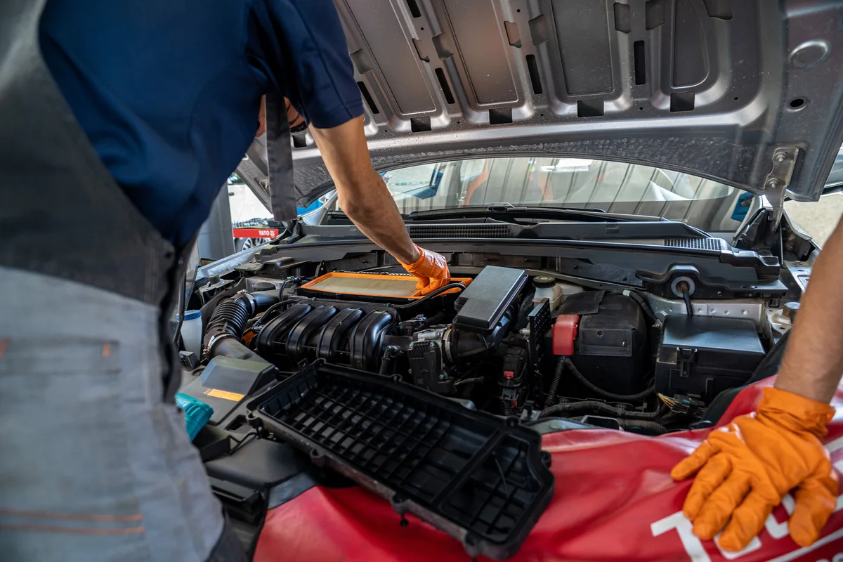 Mecánico con guantes naranjas revisa un filtro de aire dentro del compartimento del motor de un automóvil con el capó abierto.