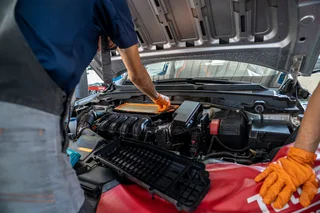 Mecánico con guantes naranjas revisa un filtro de aire dentro del compartimento del motor de un automóvil con el capó abierto.