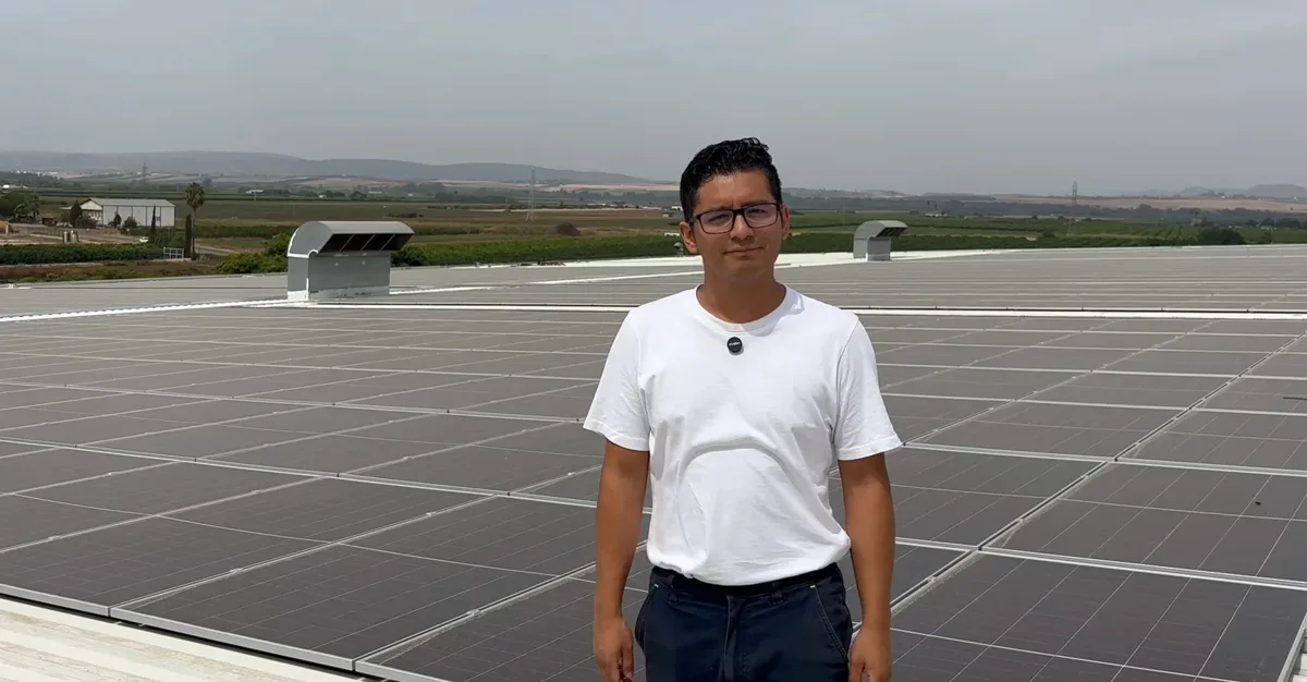 Un hombre con camiseta blanca y gafas delante de una extensa instalación fotovoltaica sobre la cubierta de una nave industrial.