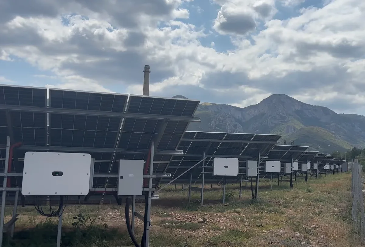 Filas de paneles solares montados sobre estructuras metálicas, con equipos eléctricos blancos bajo los módulos y montañas al fondo.